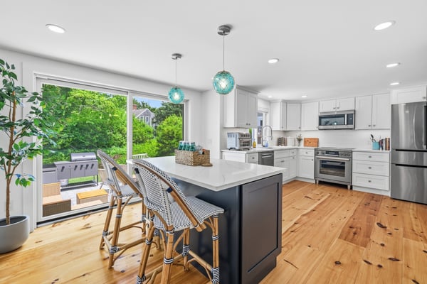 Modern kitchen with white cabinetry, stainless steel appliances, green subway tile backsplash, wooden round table, and large windows overlooking trees.
